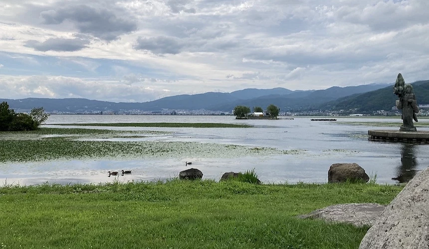 綠色草坪與灰色石雕像靠近湖畔,蔚藍湖水倒映城鎮與遠景山景,鴨隻游泳於淺灘,層疊青山環繞遠景,白色蓬鬆雲朵與淡藍天空,日本湖景風景秀麗宜人。