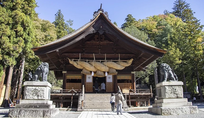 褐色木造神社正殿,翹角屋頂與金色裝飾,大麻繩與稻草飾物懸掛,石獅雕像守衛兩側,木製階梯與白色欄杆,參拜者攀登,高聳綠樹環繞,晴朗藍天映照,日本古神社建築莊嚴宏偉。