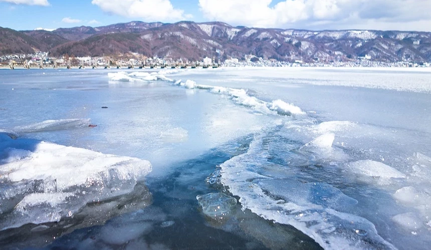 冰凍湖面漂浮白色冰塊,蔚藍湖水與冰層相間,背後城鎮建築靠近岸邊,層疊褐色與白色雪山環繞遠景,白色蓬鬆雲朵與深藍天空,日本冬季湖景冰凍壯闊秀麗。