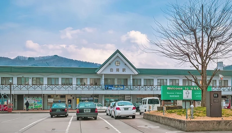白楬綠屋頂車站建築,寫有日文標語與歡迎牌,停車場與山林景觀映襯,日本鄉間火車站。