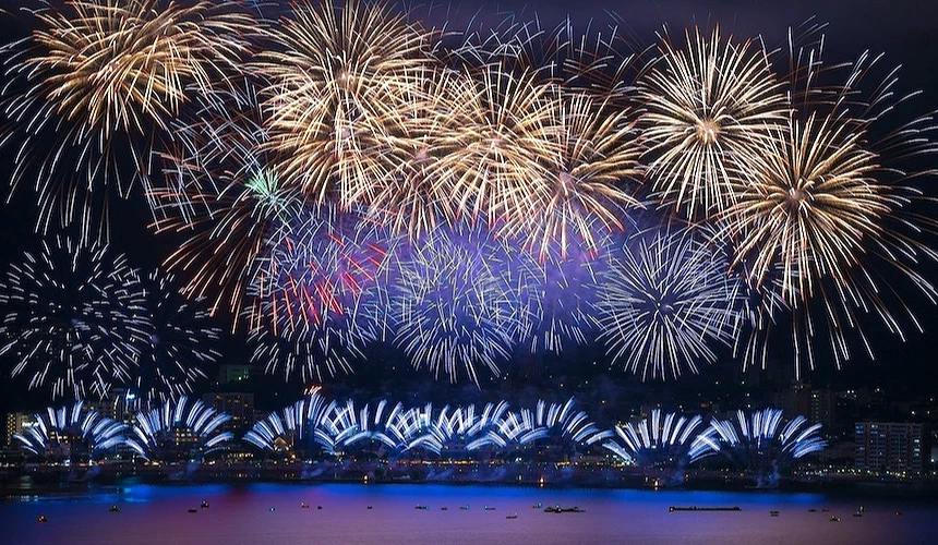 金色與藍色煙火在夜空綻放,紫紅色光芒交織,湖面倒映藍色水幕與城鎮燈火,小舟散佈水面,暗藍夜空映照,日本湖畔盛大煙火祭典壯觀絢麗。
