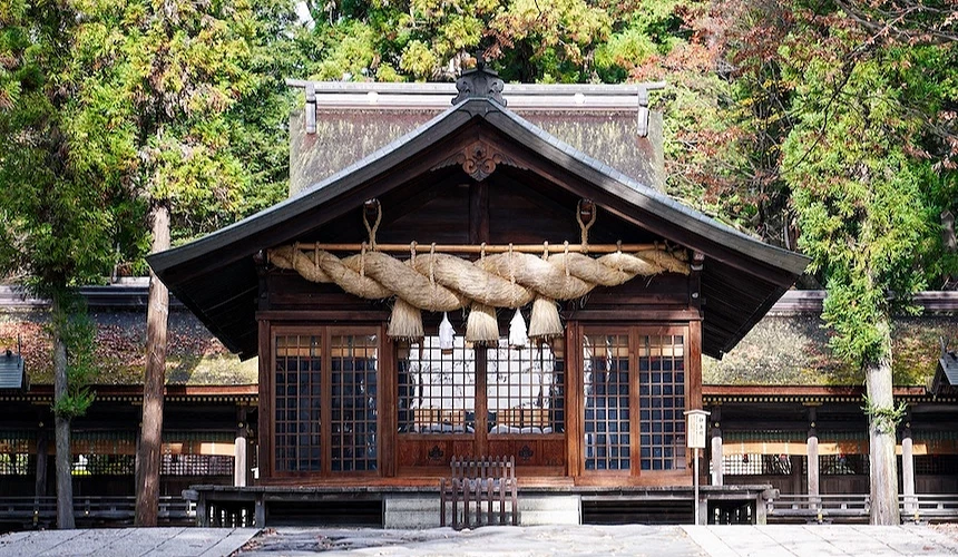 褐色木造神社殿堂,黑色翹角屋頂與白色牆體,大麻繩與稻草飾物懸掛,紅褐色木製拉門與黑色格子窗,白紙符咒懸掛,木製平台與階梯,灰色石牆基座,高聳綠樹與紅楓環繞,日本古神社建築精緻莊嚴。