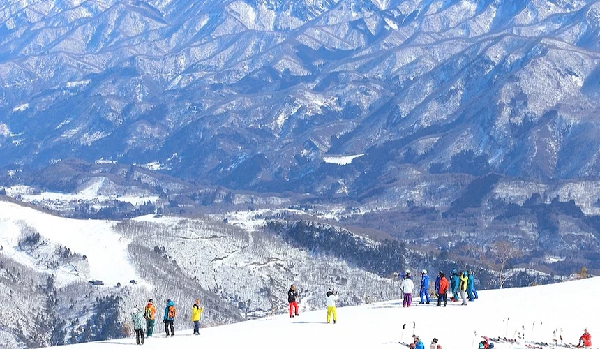 滑雪場白雪皚皚,遊客穿著彩色滑雪衣分散山坡,遠景層疊雪山與裸露褐色山脈延伸,晴朗藍天映照日本冬日滑雪勝地壯麗風景。