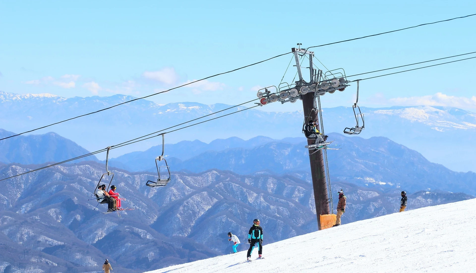 滑雪纜車運行山坡,遊客乘坐椅子上升,白雪覆蓋前景與滑雪者散落,層疊藍色山脈與晴朗天空延伸遠景,日本高山滑雪度假村風景秀麗。