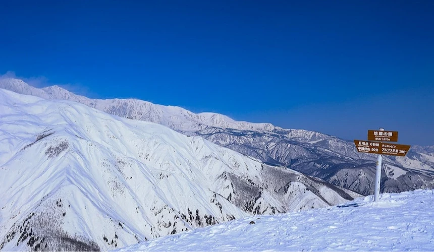 山頂木製指示牌標示日文站名與方向,白雪覆蓋山坡與褐色裸露灌木,層疊雪山延伸遠景,晴朗深藍天空映照日本高山滑雪區壯闊風景。
