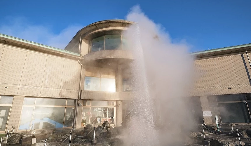 溫泉蒸汽柱升騰現代建築前,米色磚牆與綠色屋頂,圓形玻璃窗與木製欄杆,遊客沐浴溫泉,白色蒸汽繚繞,晴朗藍天映照,日本溫泉設施蒸汽騰騰特色獨特。