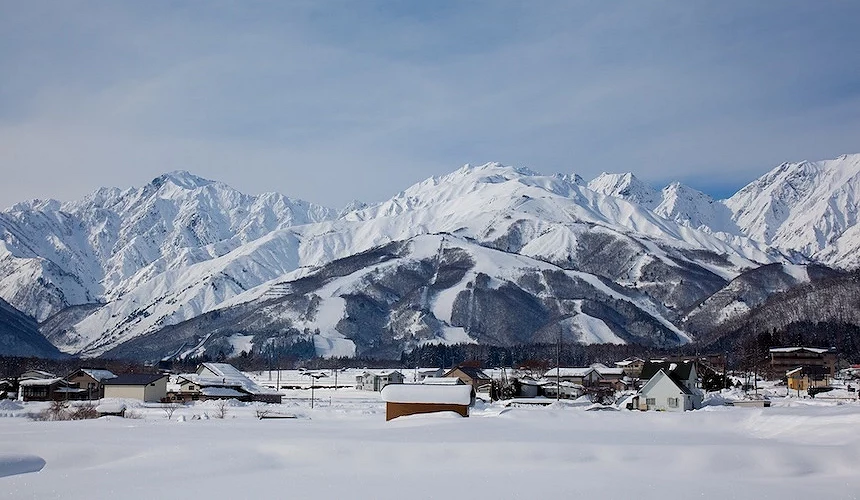 雪白湖面與鄉村房舍靠近,背後層疊雪山聳立,褐色裸露山坡與白雪相間,晴朗藍天映照日本北方冬日鄉間風景寧靜壯麗。