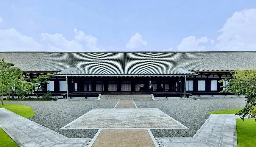 黑色木造寺院建築對稱排列,灰色瓦片屋頂與綠色邊飾,白紙拉門與木製柱子整齊,碎石廣場與綠草坪對稱鋪設,石板通路中央,晴朗藍天映照日式古寺建築莊嚴宏偉。