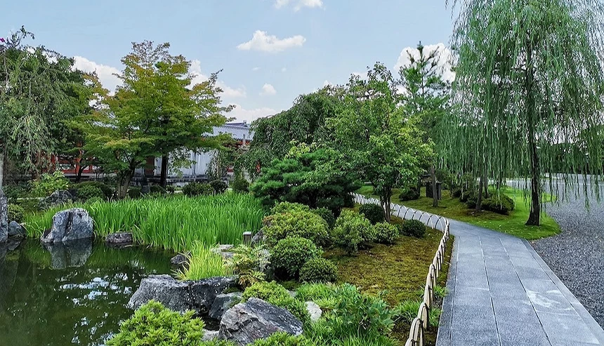 石板步道蜿蜒庭園,清池倒映綠樹,垂柳與松樹聳立,灰石與綠灌木點綴,白色木製柵欄環繞,遠景寺院建築與藍天映襯,日式禪意庭園清幽雅致。
