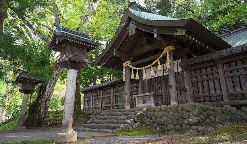 褐色木造神社殿堂,藍灰色翹角屋頂,大麻繩與稻草飾物懸掛,白紙拉門與黑色木製欄杆,石製燈籠與木製燈籠排列,灰色石堆砌基座,高聳綠樹環繞遮蔭,石板參道鋪設,日本古神社建築歷史深遠莊嚴。