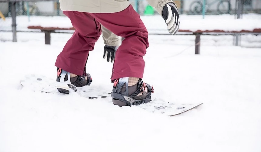 滑雪者穿著紅褐色滑雪褲與白上衣,腳踩滑雪板與黑色靴子,雙手戴黑色手套,白雪地面與背景木製休息區模糊,冬季滑雪運動特寫。