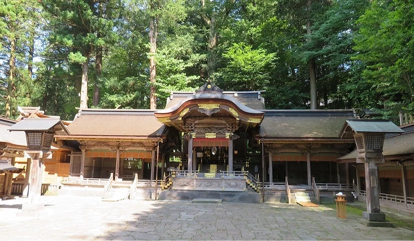 木造寺院正殿,金色邊飾與翹角屋頂,側廊與木製欄杆環繞,石板廣場寬敞,高聳綠樹背景,日式古刹建築莊嚴宏偉。