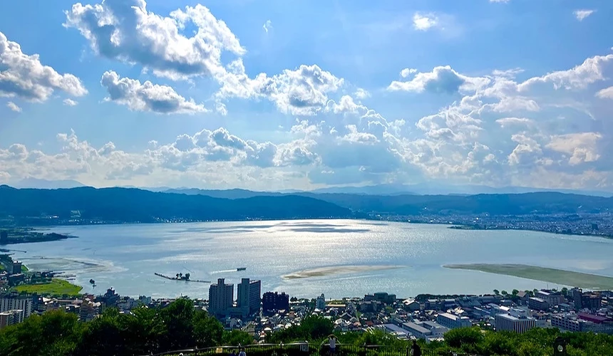 蔚藍寬闊湖面倒映白雲,城鎮與綠樹靠近岸邊,層疊青山環繞遠景,白色蓬鬆雲朵與深藍天空,湖景壯闊秀麗。