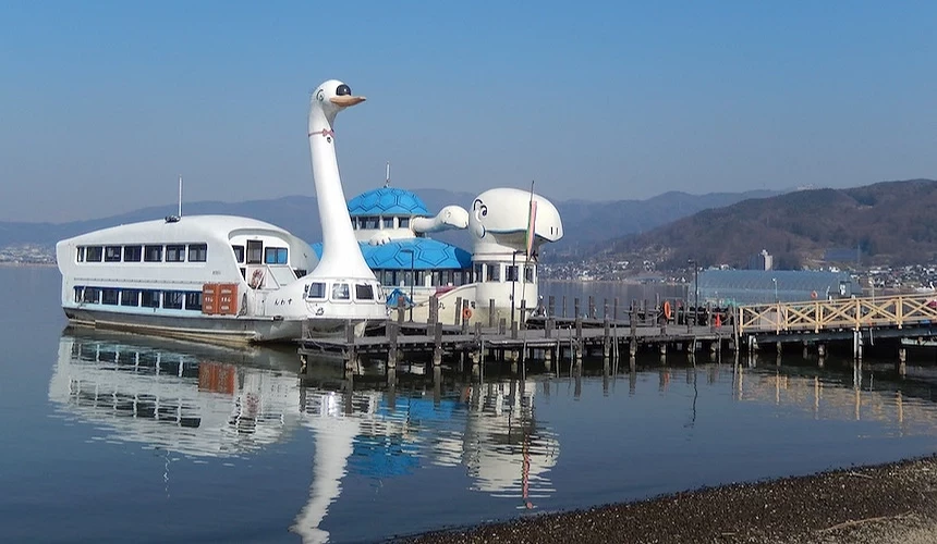 白色鵝形遊船停靠木製棧橋,藍色屋頂與白色船身,長頸雕像聳立船頭,倒映平靜湖面,背後層疊青山與城鎮靠岸,晴朗藍天映照,日本湖畔特色觀光遊船趣味獨特。
