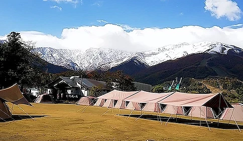 露營帳篷與篝火環繞草坪,粉紅帳篷與黃色天幕搭建,背後層疊雪山聳立,綠林與褐色山坡點綴,晴朗藍白雲天映照日本高山露營地壯麗。