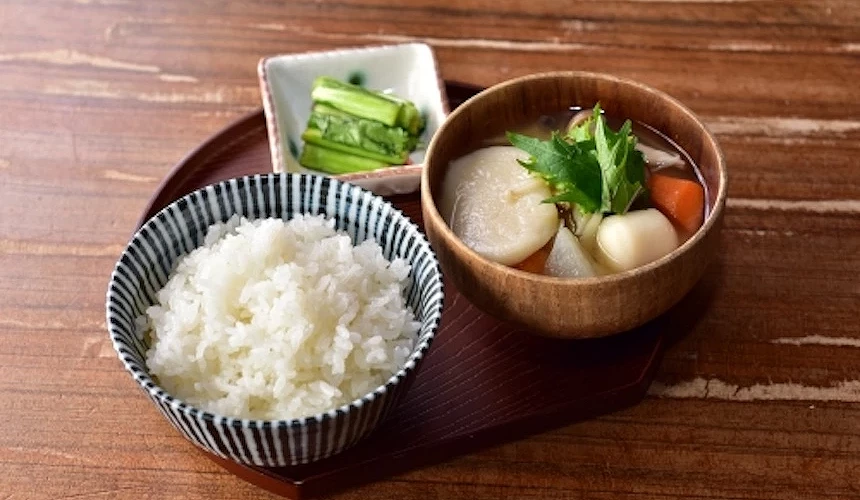 木製餐盤盛裝白飯與木碗湯品,湯內盛白色蘿蔔與紅蘿蔔,綠葉點綴,白瓷碟盛小黃瓜,日式簡單定食精緻。