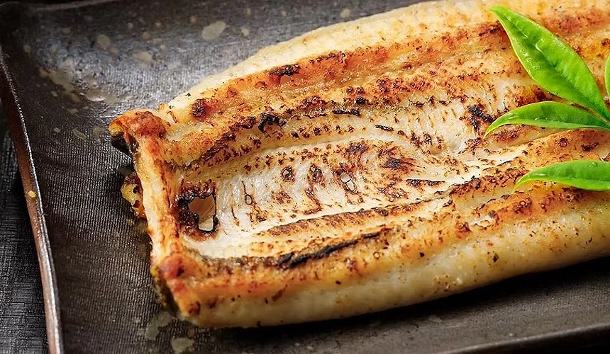 黑色陶盤盛裝烤鰻魚,表面焦香金黃與褐色紋理,綠色豆莢點綴,黑色筷子與木製筷架,日本烤鰻魚料理精緻誘人。