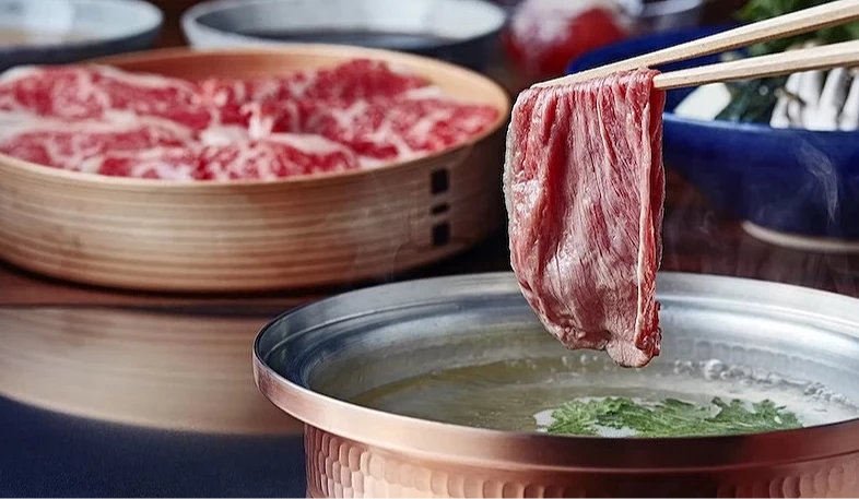 筷子夾起紅肉片浸入高湯鍋,竹籃盛裝肉片與配菜,白瓷碗與藍色碟盤整齊,日式火鍋用餐情景。