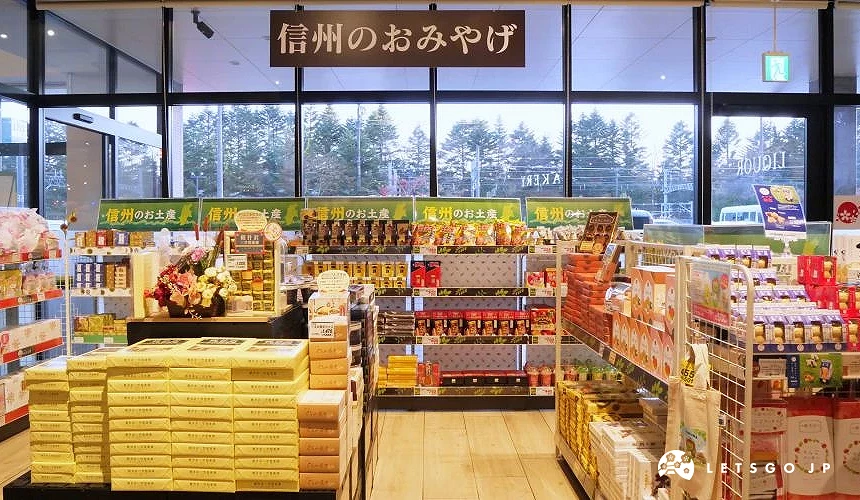 寬敞特產店內部,黃色包裝禮盒堆疊展示櫃,日文標語與商品架排列整齊,玻璃窗外綠林景觀,日本信州土產紀念品店寬闊明亮。