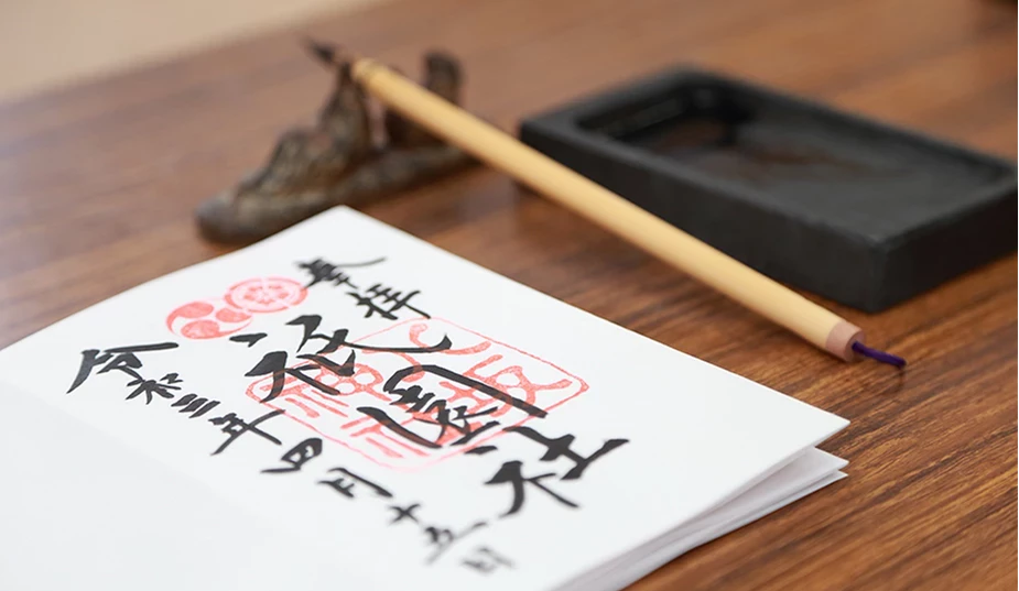 木桌上擺放著一本打開的手寫御朱印帳,上方有墨書毛筆字與朱紅色印章,背景搭配著毛筆、筆架與硯台。