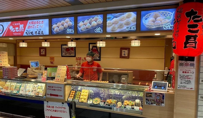 日式丸子店鋪寫有日文標語,螢幕展示食物照片,玻璃櫃台盛裝彩色丸子,紅色燈籠點綴典雅。