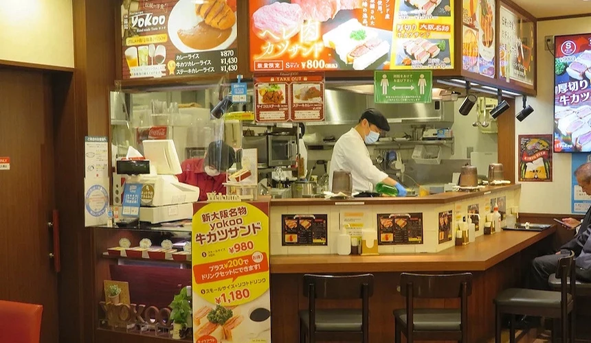 日式便當店內部,發光菜單展示食物與價格,廚師操作烹飪,木質吧台與紅色座椅整潔。