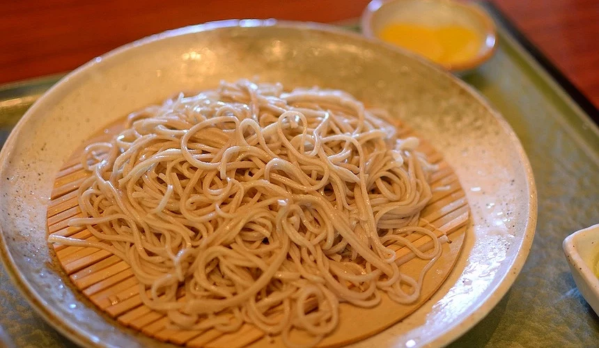 褐色陶碗盛裝棕色湯汁與蕎麥麵,麵條蜷曲堆疊,背景玻璃杯盛黃色湯汁,日式溫蕎麥湯麵精緻美味。