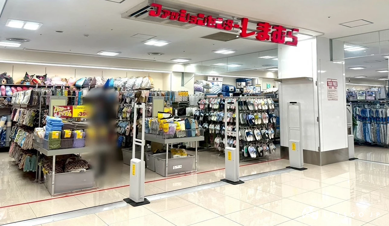 商場內掛有紅色「しまむら」日文與英文字樣招牌的店面入口,前方設有防盜感應門與生活雜貨商品陳列架。