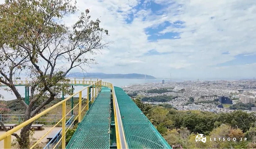 山頂滑梯與城市全景,黃綠色金屬滑梯欄杆,老樹與山林,遠景城市建築與海灣,遠方島嶼與山脈,晴朗藍天白雲,遊樂園景觀壯闊。