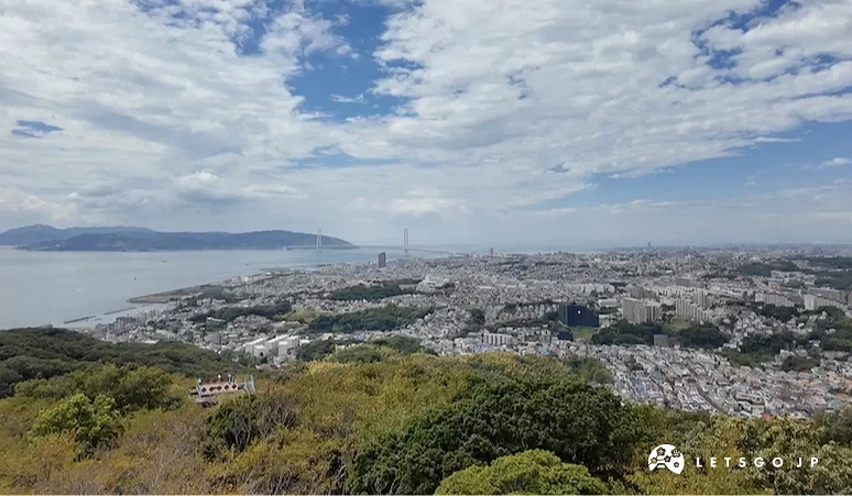 山頂俯瞰城市全景,灰色城市建築密集,藍色海灣與遠景島嶼,綠樹山林環繞前景,白色雲朵與藍天,日本濱海城市壯闊景觀。