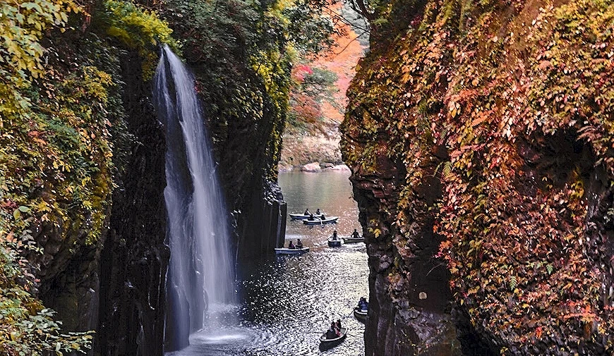 峽谷河道中多艘遊客划船,左側有瀑布落下,兩岸岩壁覆滿秋季紅葉