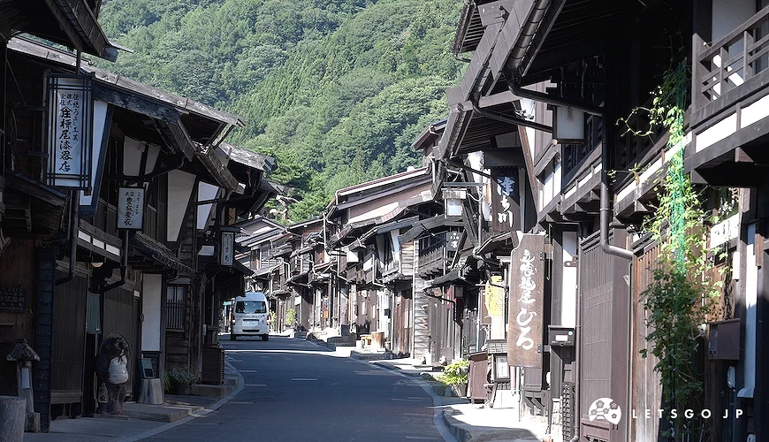兩側黑木造町家林立的街道,遠處綠山,一輛白色廂型車行駛其中,左側招牌寫有「金物屋漆器店」
