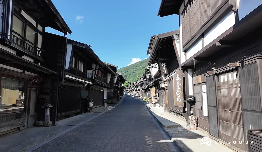 晴天下兩側黑色木造町家延伸的街道,右側寫有日文招牌,遠景可見綠山