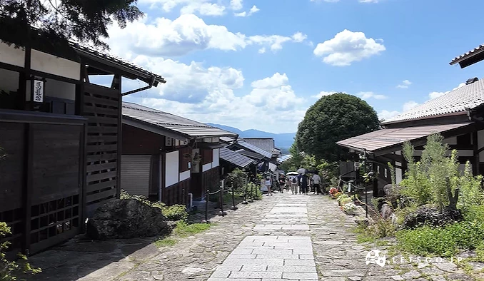 石板小路兩側為傳統木造建築,遠方可見山脈與積雲,數名行人走於路上