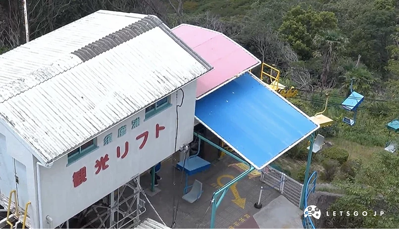 索道站房與平台,白色建築與紅字日文標示,粉紅藍色遮陽棚,黃色安全欄杆,綠樹山林環繞,山頂纜車站設施。