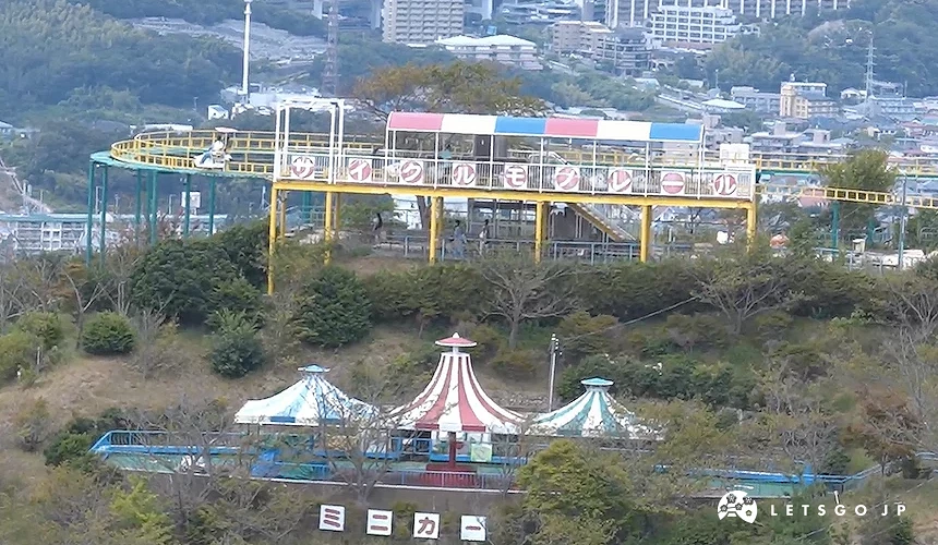 遊樂園俯瞰全景,彩色旋轉木馬與藍紅白帳篷,黃色與綠色遊樂設施,綠樹山林環繞,遠景城市建築與高樓,晴朗藍天映照,山頂遊樂園設施完整。