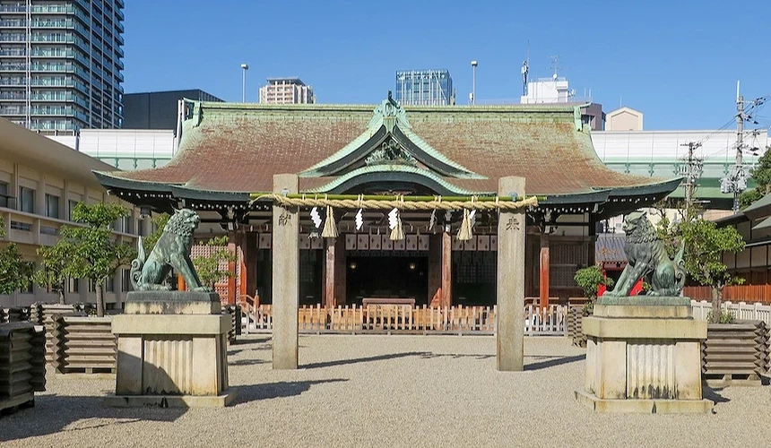 日本神社主殿,綠色銅瓦屋頂與棕色木質結構,石獅守護兩側,白色木欄杆與紅色燈籠,砂礫參道,背景現代高樓,傳統與當代建築融合。