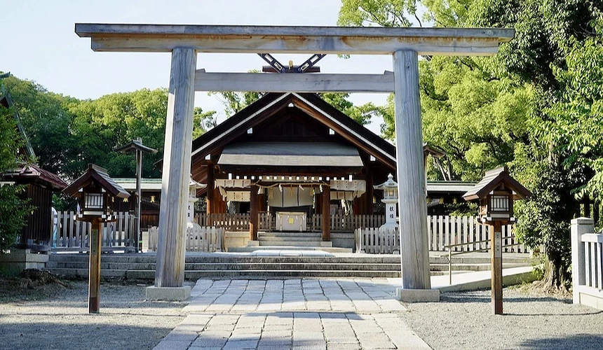 日本神社入口,灰色木製鳥居與石板參道,深棕色主殿與白色木欄杆,翠綠樹木環繞,晴朗藍天映照,傳統神聖建築風格。