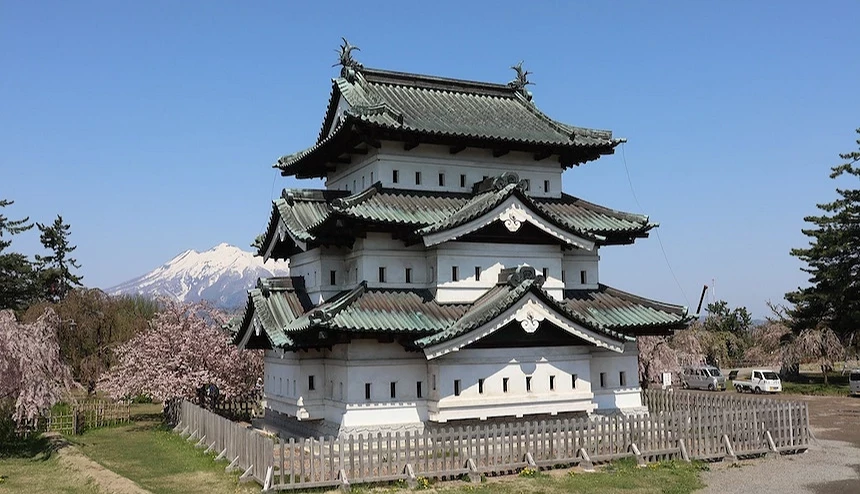 日本古城堡,灰瓦翹角屋頂與綠色邊飾,白色石牆與木製欄杆,背後雪山聳立,粉紅櫻花樹環繞,晴朗藍天映照,春季古城風景壯麗。