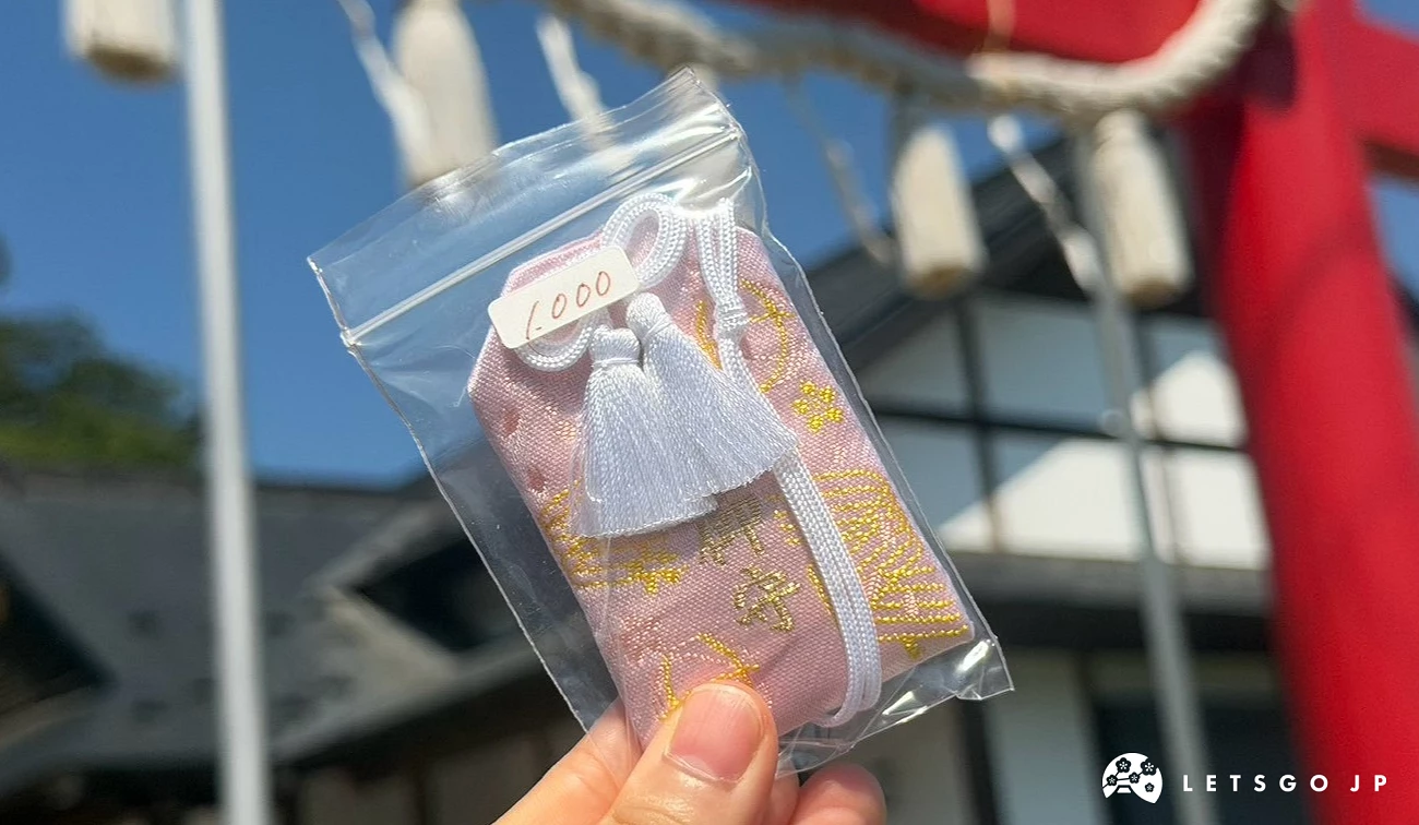 手中拿著透明封套包裝的「愛子御守」,淡粉色布面上繡有精緻金線圖案與白色流蘇。