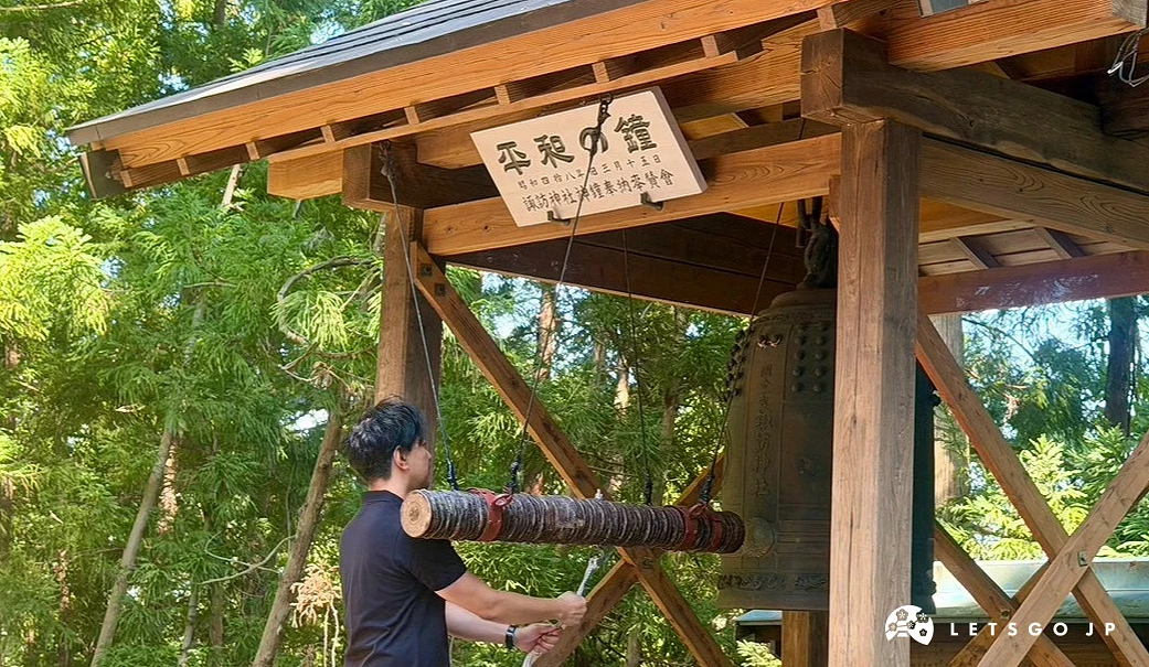 一名男子站在木造鐘樓下,雙手握著撞木準備敲響大型古銅鐘,背景為一片綠意盎然的樹林。