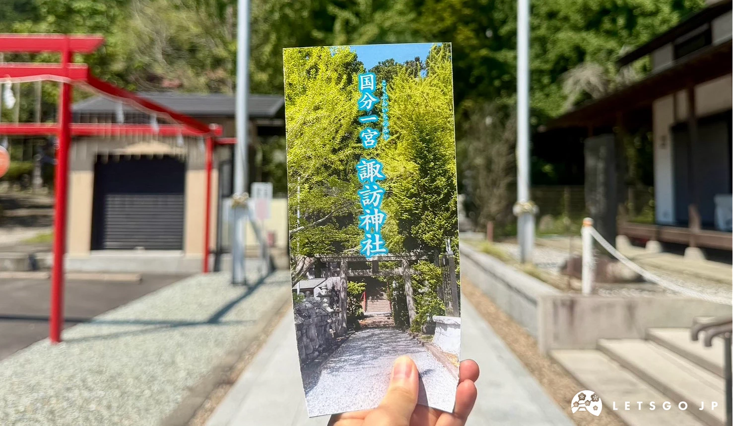 手中拿著直式設計的神社參拜簡介摺頁,上面印有藍天下的神社鳥居照片,背景為神社石板路與綠樹。