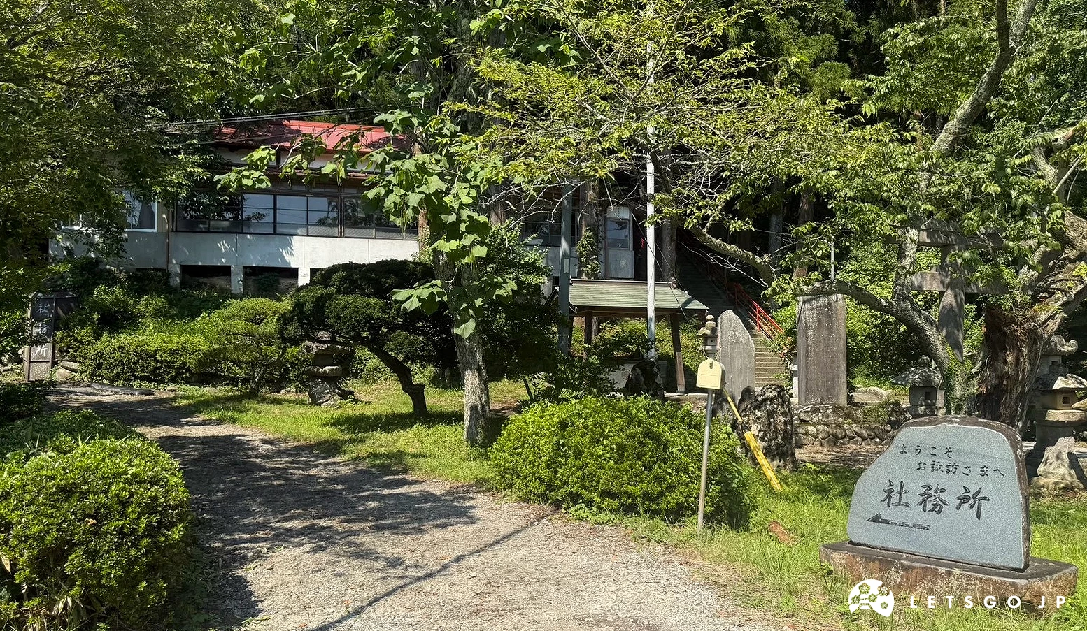 國分一宮諏訪神社境內綠意盎然的樹林與修剪整齊的圓形灌木,旁邊立有刻著社務所的石碑,陽光透光樹葉灑在地面。