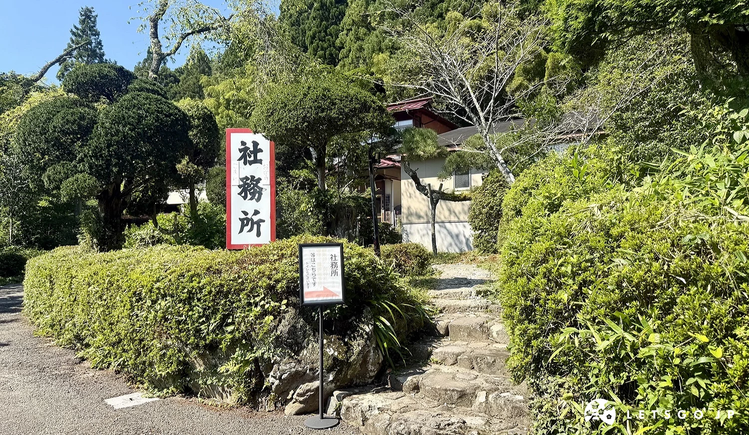 道路旁修剪整齊的綠色灌木叢,上方樹立著白底紅邊寫有黑色「社務所」字樣的立牌,右手邊有一條通往後方山林建築的石階小路。