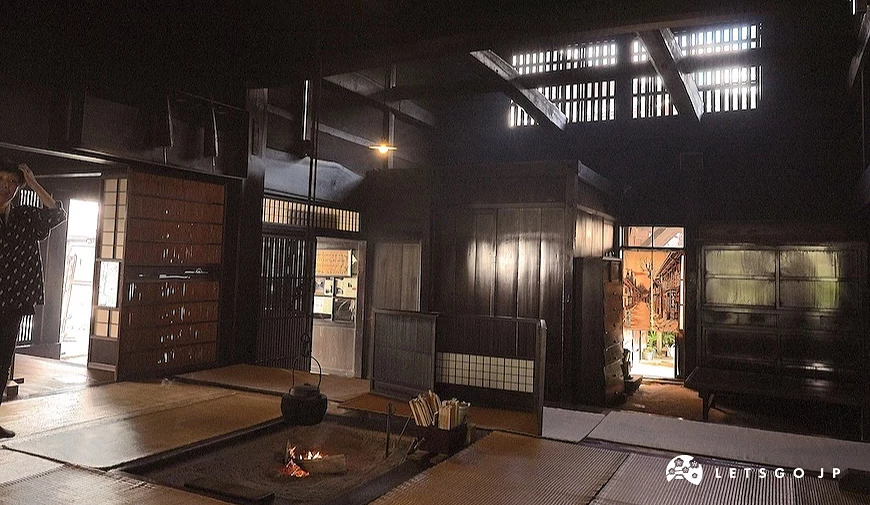 夜間室內場景,黑色木造建築內可見地爐火光、柴薪與榻榻米地板,背景有多扇木門與格柵窗透光