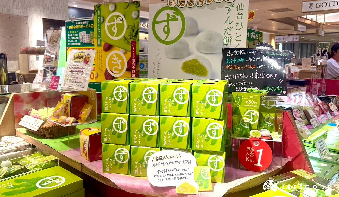 伴手禮專櫃陳列的毛豆泥餅乾與特產禮盒,鮮綠色包裝堆疊整齊,後方有毛豆泥麻糬的海報展示。