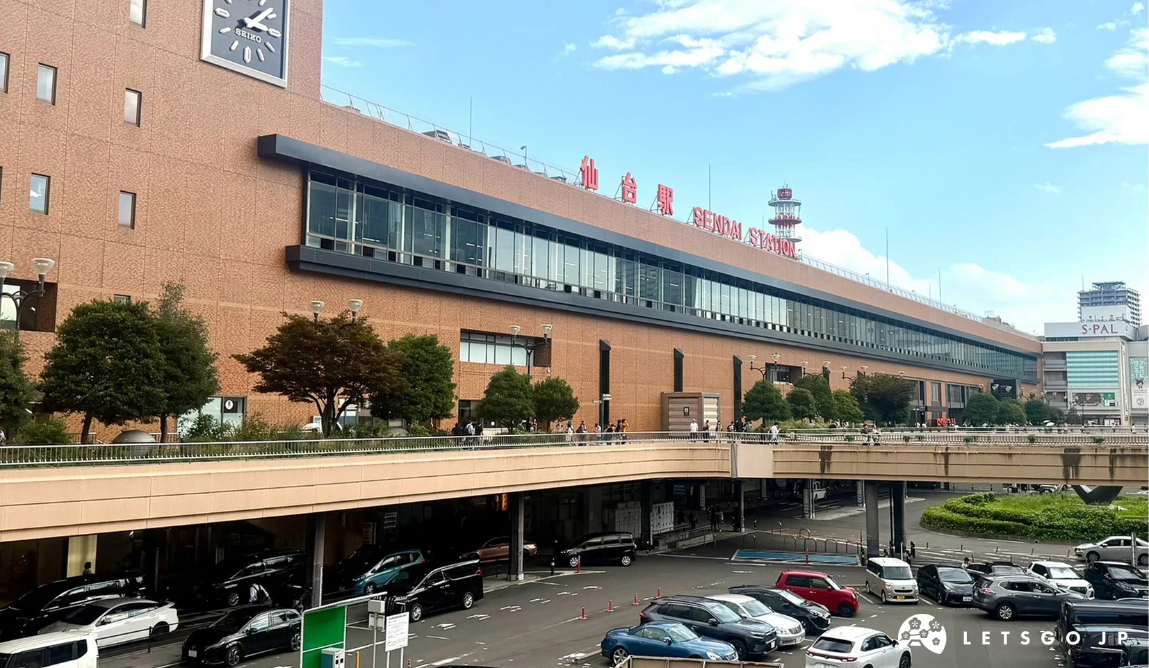 晴朗藍天下的 JR 仙台車站大樓外觀,紅磚牆面頂端懸掛紅色「仙台駅」字樣標誌,前方為寬廣的高架行人天橋與車道。
