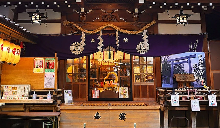 神社商店,深棕色木質建築與翹起屋頂,金色圓形裝飾,白色暖簾與繩結掛飾,木製櫃檯與玻璃窗展示商品,黃色燈籠,翠綠樹木背景,傳統日式風格。