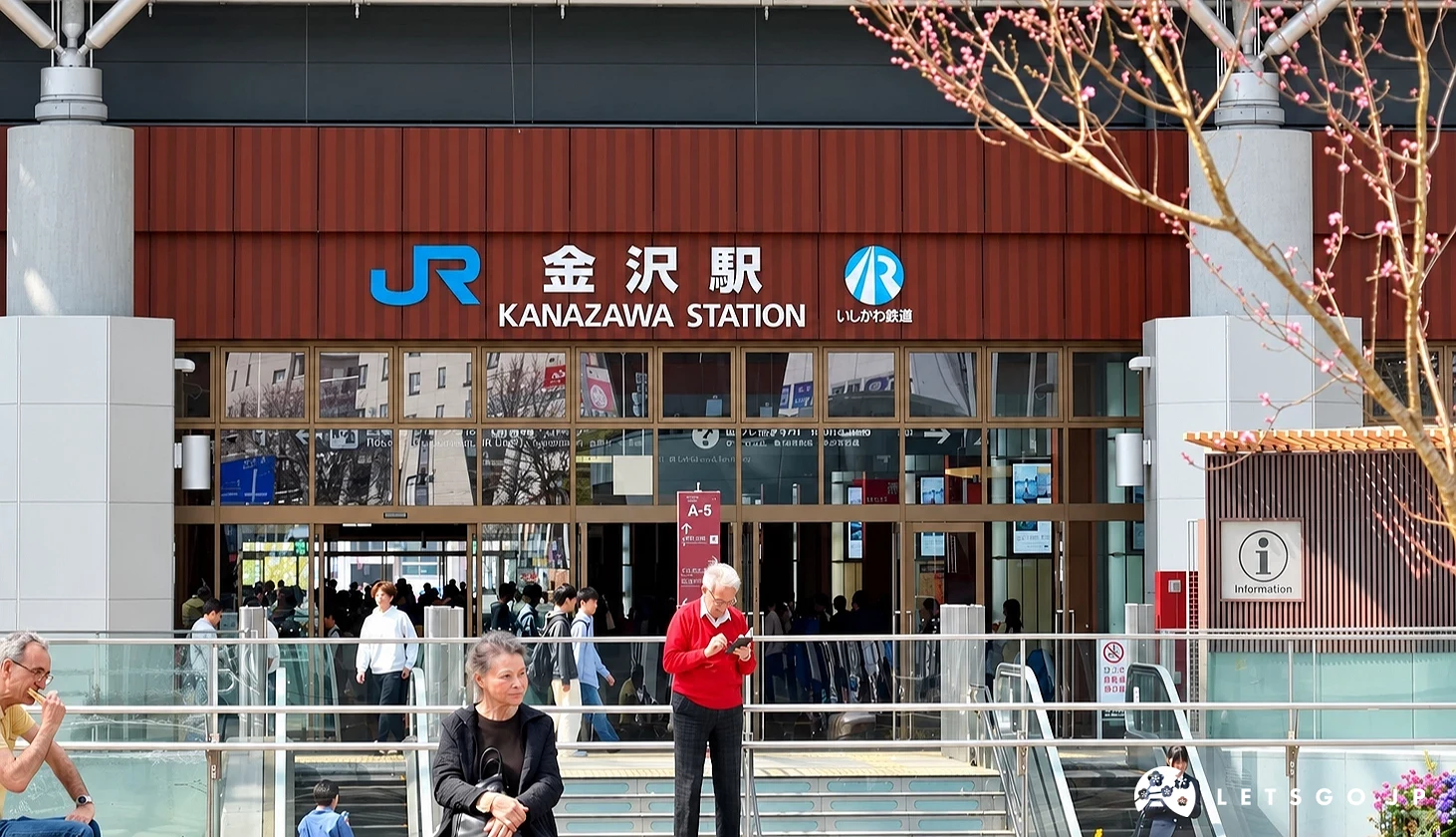 金澤車站入口上方寫有「金沢駅」與「KANAZAWA STATION」標示JR與石川鐵道,前方有行人,右側有粉色花朵枝條