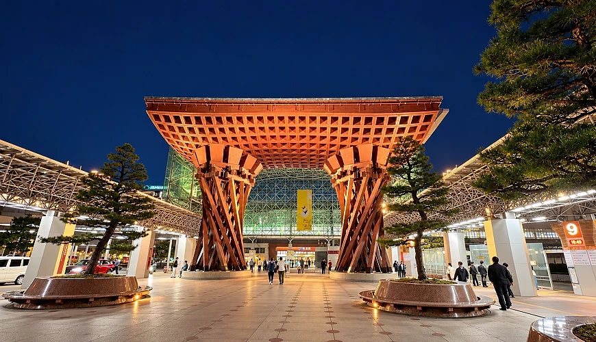 夜間木造鼓門與玻璃穹頂建築前的廣場上有行人與盆栽松樹
